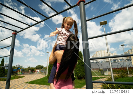 Mother lifts her daughter in a sunny playground on a summer afternoon 117458485