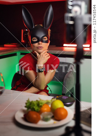 A beautiful girl in a bdsm-style rabbit mask and a bright red dress is chatting sweetly in a stream in the kitchen in neon light 117458544