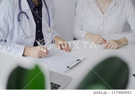Doctor and a patient. The female physician, wearing a white medical coat over a dark blue dotted blouse, using clipboard and making notes during a consultation in the clinic. Medicine concept 117460042