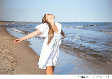 Happy smiling beautiful woman is on the ocean beach in a white summer dress, open arms 117460061