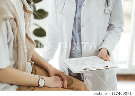 Doctor and patient in clinic. Friendly physician explaining something to young woman while keeping a clipboard with medical records. Medicine concept 117460071