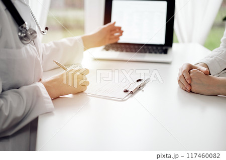 Doctor and patient sitting and discussing something near each other at the white desk in clinic. Female physician is pointing into laptop screen. Medicine concept 117460082