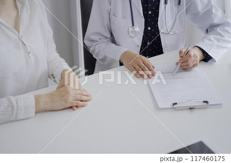 Doctor and a patient. The female physician, wearing a white medical coat over a dark blue dotted blouse, using clipboard and making notes during a consultation in the clinic, close up. Medicine 117460175