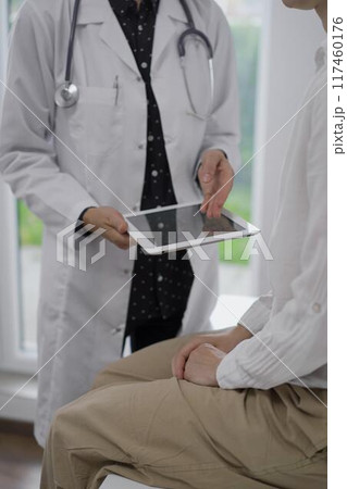 Doctor and a patient discussing something. The female physician is pointing into tablet computer screen with a medical notes besides a young woman during a consultation in the clinic Doctor and a patient discussing something. The female physician is pointing into tablet computer screen with a medical notes besides a young woman during a consultation in the clinic 117460176