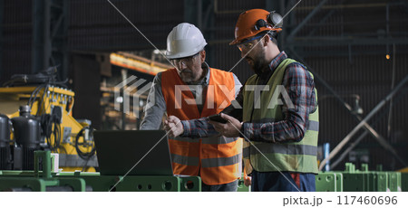 Two heavy industry engineers work using laptop and mobile phone at factory Two heavy industry engineers work using laptop and mobile phone at factory 117460696