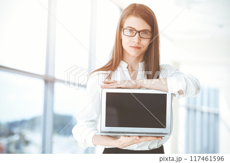 Beautiful female specialist with laptop computer standing in sunny office and smiling charmingly. Business people concept 117461596