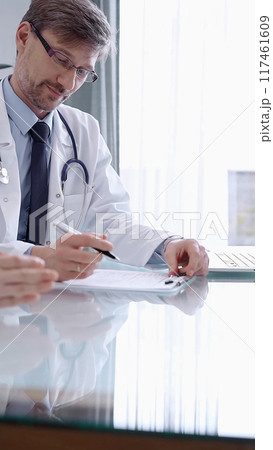 Doctor consulting patient. Male physician is pointing into medical records on a clipboard while talking with a woman in a clinic, close up. Medicine concept 117461609