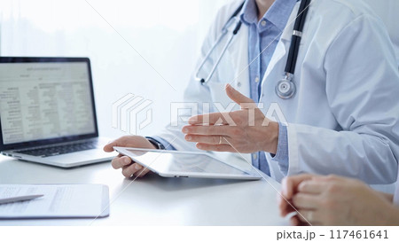 Doctor and a patient. The physician, wearing a white medical coat over a blue shirt, is using tablet computer during a consultation in the clinic. Medicine and science 117461641