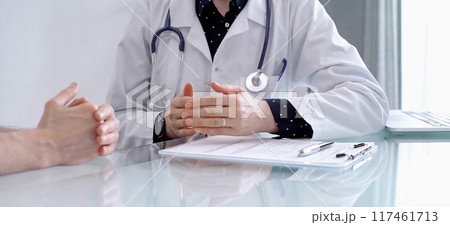 Doctor consulting patient. Female physician is gesturing with his hands over a glass desk while talking with a man in a clinic. Medicine concept Doctor consulting patient. Female physician is gesturing with his hands over a glass desk while talking with a man in a clinic. Medicine concept 117461713
