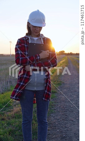 Woman engineer is standing straight and taking notes with a clipboard in a field with wind turbines, as the sun sets. Clean energy and engineering concept Woman engineer is standing straight and taking notes with a clipboard in a field with wind turbines, as the sun sets. Clean energy and engineering concept 117461715