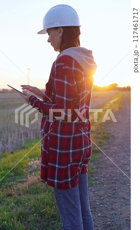 Woman engineer wearing a white protective helmet is taking notes with a clipboard in a field with wind turbines, as the sun sets. Clean energy and engineering concept 117461717