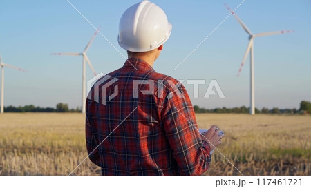 Man engineer at work is wearing a white protective helmet and taking notes with a clipboard in a field with wind turbines, as the sun sets. Clean energy concept Man engineer at work is wearing a white protective helmet and taking notes with a clipboard in a field with wind turbines, as the sun sets. Clean energy concept 117461721