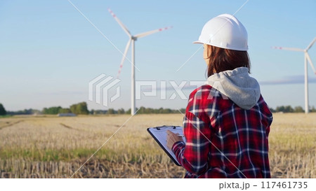 Female engineer wearing a white protective helmet is taking notes with a clipboard in a field with wind turbines, as the sun sets. Clean energy concept Female engineer wearing a white protective helmet is taking notes with a clipboard in a field with wind turbines, as the sun sets. Clean energy concept 117461735
