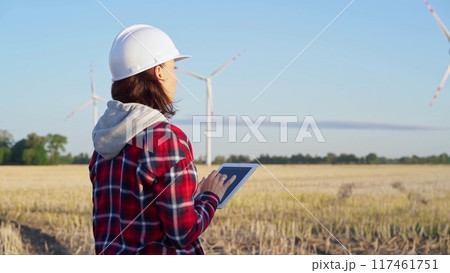 Woman engineer wearing a white protective helmet is taking notes with a tablet computer on a field with wind turbines, as the sun sets. Clean energy and engineering concept 117461751