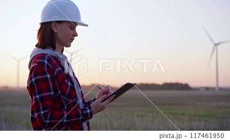 Female engineer taking notes on a clipboard on a field with wind turbines, as the sun sets. Concept of clean energy and engineering audit 117461950