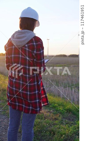 Woman engineer wearing a white protective helmet is taking notes with a clipboard in a field with wind turbines, as the sun sets. Clean energy and engineering concept 117461951