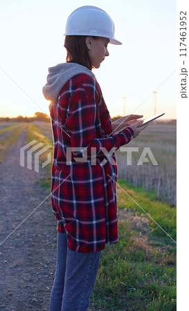 Woman engineer wearing a white protective helmet is taking notes with a clipboard in a field with wind turbines, as the sun sets. Clean energy and engineering concept 117461952