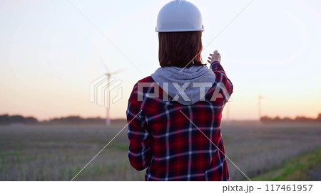 Woman engineer taking notes on a clipboard on a field with wind turbines, as the sun sets, back view. Clean energy and engineering concept 117461957