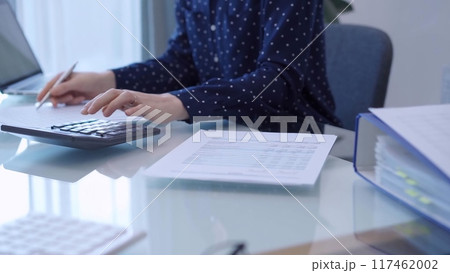 A female accountant with blue dotted blousy is using a laptop computer and calculator to calculate taxes at a glass desk in the office. Left side view A female accountant with blue dotted blousy is using a laptop computer and calculator to calculate taxes at a glass desk in the office. Left side view 117462002