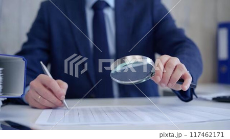 Businessman is carefully examining financial documents with a magnifying glass, highlighting the importance of thorough analysis in business. Audit and taxes 117462171