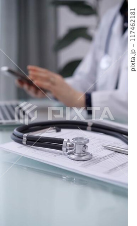 Stethoscope and medication history record form are on the glass table while doctor woman is using smartphone. Medicine theme Stethoscope and medication history record form are on the glass table while doctor woman is using smartphone. Medicine theme 117462181