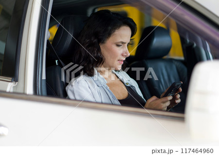 A woman is sitting in a car, looking at her phone, appearing to type or read something important on it. 117464960
