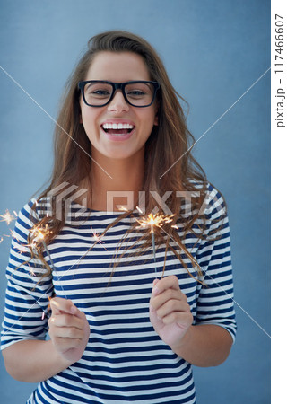 Smile, portrait and woman in studio with sparklers for celebration isolated on blue background with light. Happy, glasses and fun event with birthday or new year with glow, vision and fireworks. Smile, portrait and woman in studio with sparklers for celebration isolated on blue background with light. Happy, glasses and fun event with birthday or new year with glow, vision and fireworks. 117466607