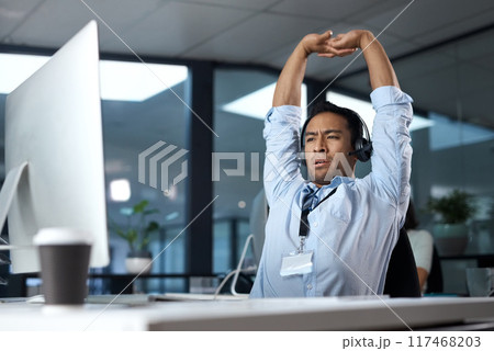 Some calls just make you question your choice in career. Shot of a young man stretching while using a headset and computer in a modern office. Some calls just make you question your choice in career. Shot of a young man stretching while using a headset and computer in a modern office. 117468203