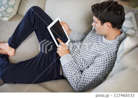 Man, tablet and relax on sofa in living room for tech, social media and research in home or indoor on weekend. Male person and looking with touch on screen for browse with internet and online 117468533