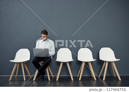 Business man, computer and waiting room of job search, opportunity or planning for interview or recruitment. Information technology candidate or worker reading on laptop and chair by wall background 117468758
