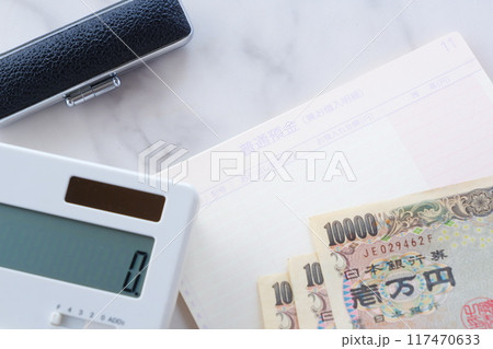 テーブルの上に開いた状態で置かれた銀行の預金通帳と、1万円札、電卓、印鑑 テーブルの上に開いた状態で置かれた銀行の預金通帳と、1万円札、電卓、印鑑 117470633