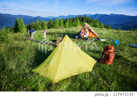 Picturesque mountain campsite on grassy hill with tents. Camper and child tend to campfire, surrounded by backpacks and gear. Rolling hills, evergreens, clear blue sky. Peaceful outdoor adventure. 117471135