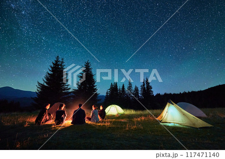 Group of friends hikers resting around campfire, surrounded by illuminated tents under stunning starry sky. Milky Way stretches above, creating magical atmosphere in peaceful mountain wilderness. Group of friends hikers resting around campfire, surrounded by illuminated tents under stunning starry sky. Milky Way stretches above, creating magical atmosphere in peaceful mountain wilderness. 117471140