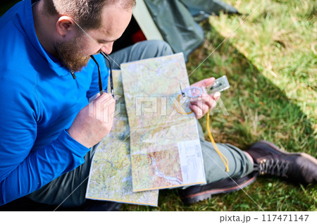 Close up man in glasses closely examines map while holding compass. He sits by his tent, deeply focused on navigation. Lush greenery, suggesting serene outdoor adventure. 117471147
