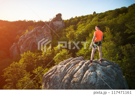 Aerial view of climber standing atop rock formation at Dovbush Rocks in Carpathian mountains, Ukraine. Sun sets, casting warm glow over lush, green forest, distant hills, creating breathtaking view. 117471161