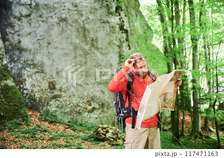 Bearded tourist man looks puzzled with surprised expression, studies map while stands in front of large rock in dense forest. Traveler with backpack and glasses, trying to figure out his direction. 117471163