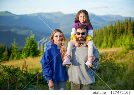 Family stands together in scenic meadow with backdrop of rolling mountains. Father carries his daughter on shoulders, both smiling and enjoying view, while mother stands beside them. 117471168