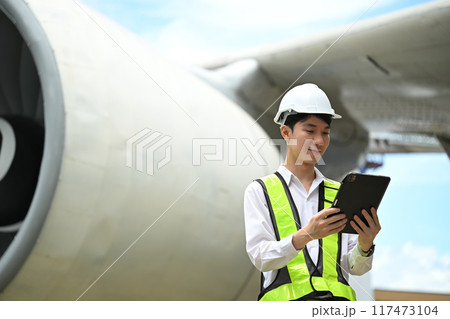Young male engineer or aviation technician using digital tablet in front of large aircraft engine 117473104