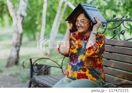 Joyful woman using her laptop as a makeshift umbrella in the park Tech-friendly environment as woman finds an unconventional use for her computer. 117473155
