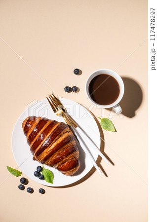 Freshly baked croissant with chocolate on a white plate and blueberries on a yellow background Freshly baked croissant with chocolate on a white plate and blueberries on a yellow background 117478297