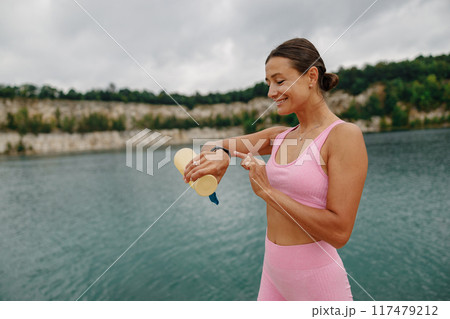 A Woman is Applying Sunscreen by the Beautiful Water While Wearing a Stylish Pink Outfit 117479212