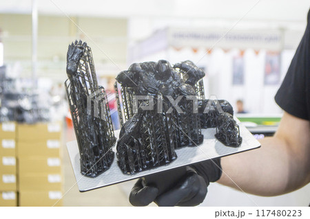 Black model with supports printed on 3D printer close-up. Man holding in hand object created on 3D printer from hardened rubber under influence of ultraviolet light. SLA 3D printing. Stereolithography Black model with supports printed on 3D printer close-up. Man holding in hand object created on 3D printer from hardened rubber under influence of ultraviolet light. SLA 3D printing. Stereolithography 117480223