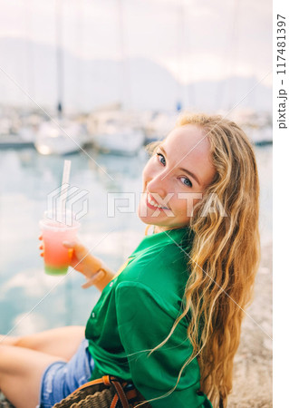 Young woman enjoying colorful bubble tea drink by seaside during sunset, with boats on background Young woman enjoying colorful bubble tea drink by seaside during sunset, with boats on background 117481397