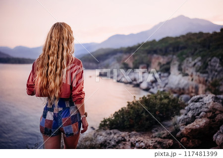 Woman gazes at sunset over a coastal landscape near mountains at dusk 117481399