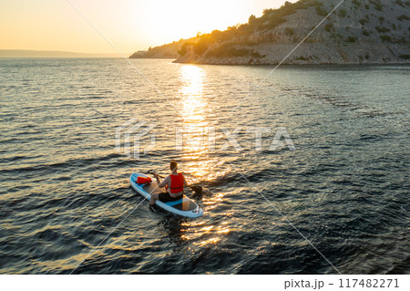 Man wearing an orange vest navigates his paddleboard through gentle waves, with the warm glow of the setting sun Man wearing an orange vest navigates his paddleboard through gentle waves, with the warm glow of the setting sun 117482271