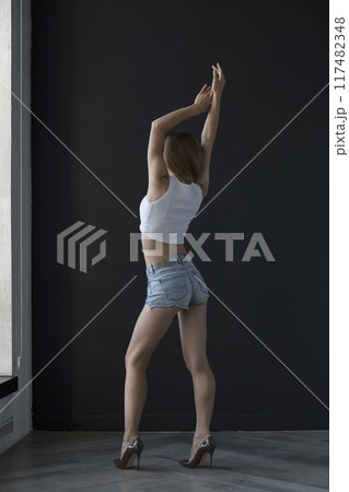 young woman in a white tank top and denim shorts on a dark background, full length portrait young woman in a white tank top and denim shorts on a dark background, full length portrait 117482348