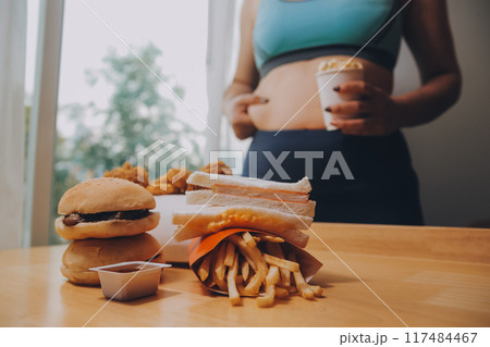 To eat or not. Cheerless plump woman looking at the burger while thinking to eat it or not 117484467