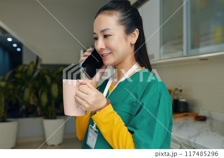 Smiling Nurse Talking on Phone While Holding Mug in Kitchen Smiling Nurse Talking on Phone While Holding Mug in Kitchen 117485296