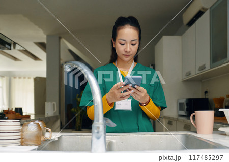 Woman Wearing Scrubs Reading from Mobile Phone in Kitchen 117485297