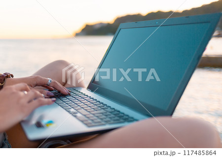 A woman works remotely on a laptop. Hands close-up. Sea in the background. The concept of freelancing and blogging 117485864